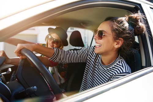 Happy woman with brown hair and sunglasses holds steering wheel and female passenger with blonde hair smiles