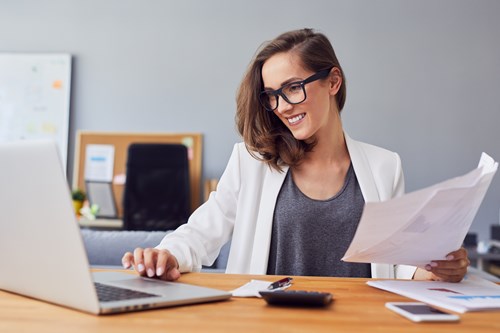 Woman with brown hair, glasses and white blazer sits at a desk typing into a silver laptop and holding sheets of paper