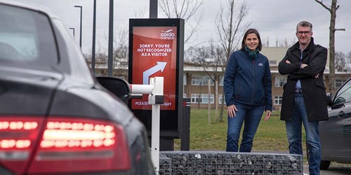 Man and woman stand behind a car park barrier being used by a car and beside a parking guidance display