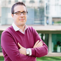 Man in white shirt and purple jumper, wears black-rimmed glasses and crosses his arms