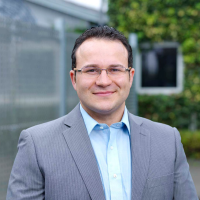 Businessmen wearing glasses, a blue shirt and grey suit jacket stands with a green background