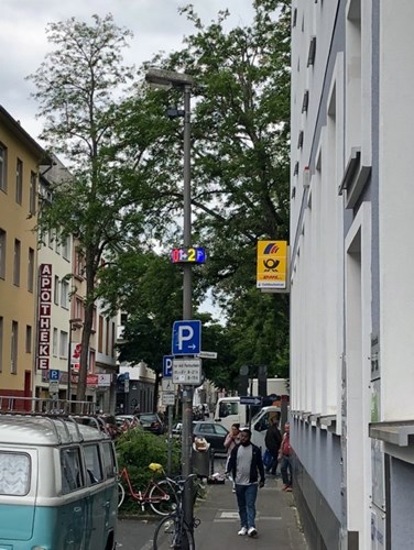 A street with lined with shops and parked cars, a lamppost has a parking guidance sign attached