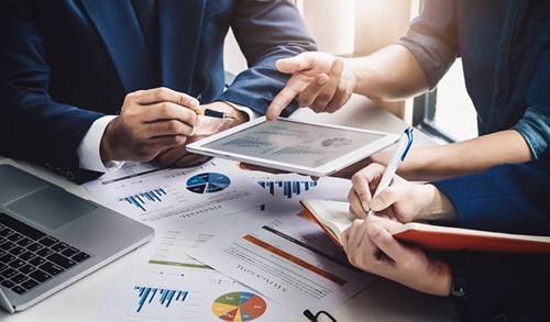 Hands of three business people sitting at a desk with a laptop, tablet, notepad and papers filled with graphs