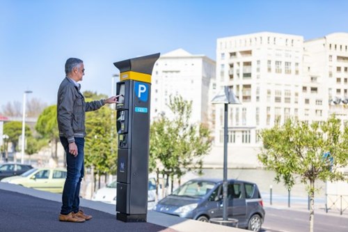 Grey-haired man in leather jacket and jeans uses a parking meter, river and bridge in background.