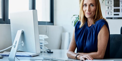 Blonde-haired business woman in blue blouse sits behind a desk and smiles at the camera