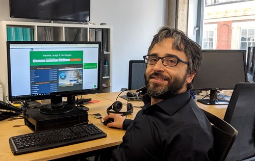 Man smiles at camera whilst sitting at desk using computer.