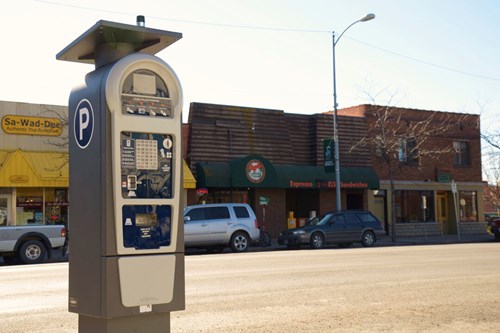 A street with shops in the background, parked cars and a parking meter in the foreground