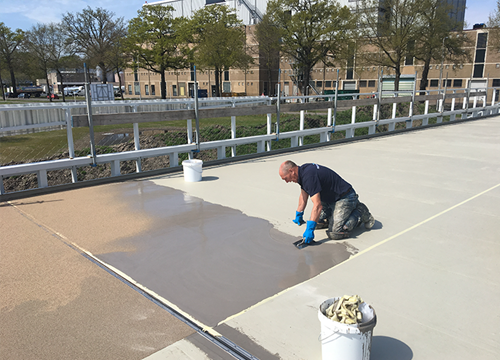 Kneeling man applies clean liquid to a concrete parking lot, trees in the background