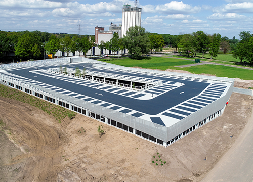 Two story parking garage sits within a building site and field with a tower block and trees in the background.